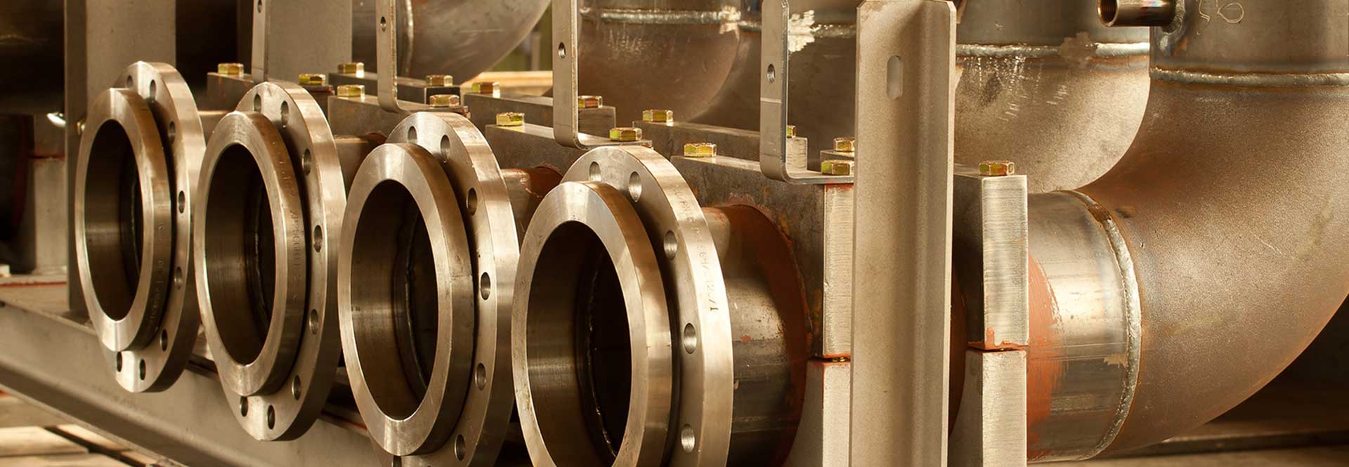 Nahaufnahme mehrerer großer metallischer Rohrflansche, die in einer Reihe angeordnet und mit Industrierohren verbunden sind - ein Beweis für die zertifizierte Fertigung und die im Apparatebau typische Präzision. Schweißmarkierungen und Bolzen befestigen jeden Flansch sicher.