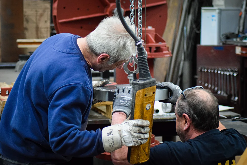 Zwei Männer mit Arbeitshandschuhen bedienen ein schweres Maschinenteil mit einem Metallkettenzug in einer auf Anlagen- und Stahlbau spezialisierten Industriewerkstatt. Ein Mann hält einen gelben Steuerkasten, während der andere bei der Positionierung hilft.