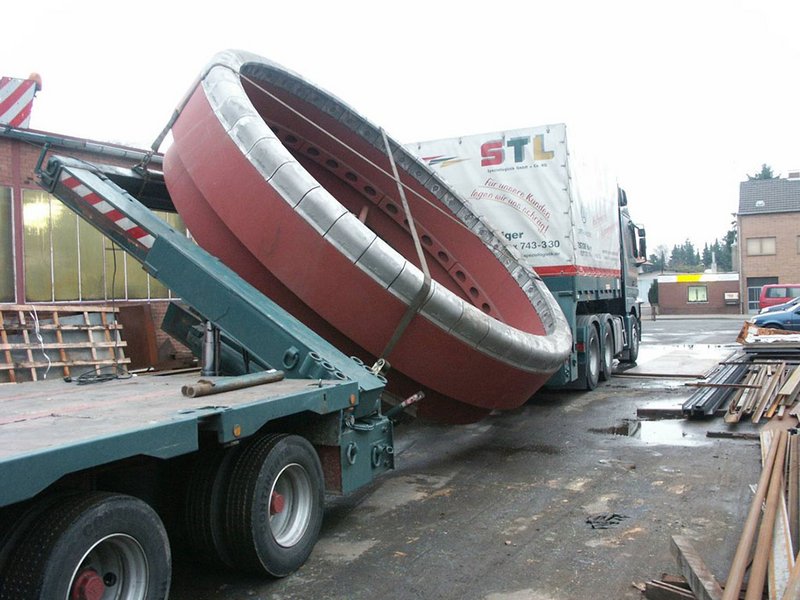 Ein großes, rundes Maschinenbau-Bauteil wird mit Hilfe eines Kippmechanismus auf einen Tieflader verladen. Der Lkw steht in einem Industriehof, der von Gebäuden und Metallmaterialien umgeben ist und die Präzision der Apparatebauarbeiten widerspiegelt.