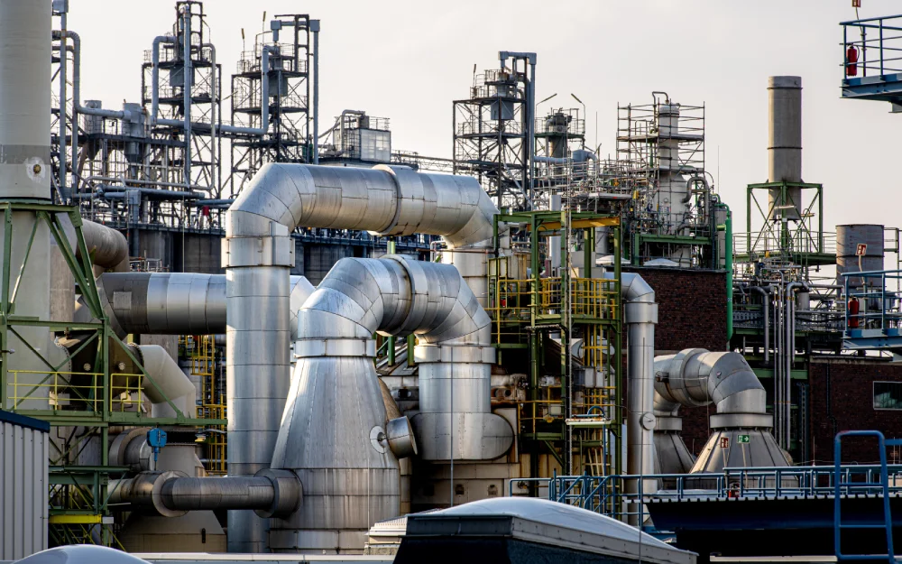 Industrieanlage mit großen Metallrohren, Tanks und komplexen Strukturen - wahrscheinlich Teil einer Chemie- oder Ölraffinerieanlage - mit Maschinenbau-Bauteilen und Stahlbau inmitten von miteinander verbundenen Maschinen und Gerüsten unter einem bewölkten Himmel.